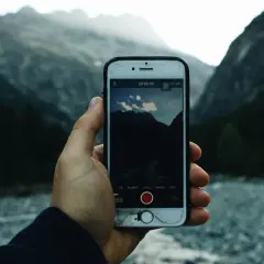 Ręka trzyma telefon z otwartą aplikacją aparatu, gotową do robienia zdjęć górskiego krajobrazu.