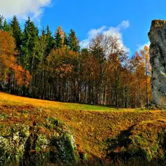 Jesienny krajobraz z wysoką skałą przypominającą maczugę Herkulesa. W tle las w barwach jesieni i błękitne niebo.