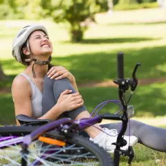 Kobieta w kasku siedzi na trawie, trzymając się za bolącą nogę. Obok leży rower. Czyżby mandat za potrącenie rowerzysty?