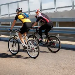 Dwóch rowerzystów jedzie obok siebie po moście. Jeden z nich macha do widza.