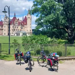 Trzy rowery z sakwami czekają przed zamkiem. W tle rozciąga się zielony park, a w oddali widać fragment rzeki Niemen.