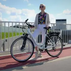Kobieta w białych spodniach i kolorowej koszuli stoi obok białego roweru elektrycznego. Czy na rower elektryczny trzeba mieć prawo jazdy?