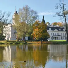 Pałac w jesiennej scenerii, otoczony drzewami o barwnych liściach, odbija się w spokojnej tafli jeziora. W oddali widać fragment puszczy bolimowskiej.