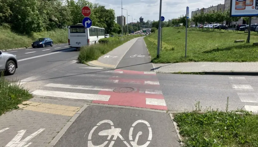 Przejście dla pieszych na ścieżce rowerowej, z oznaczeniem roweru. Autobus i samochody czekają na drodze.