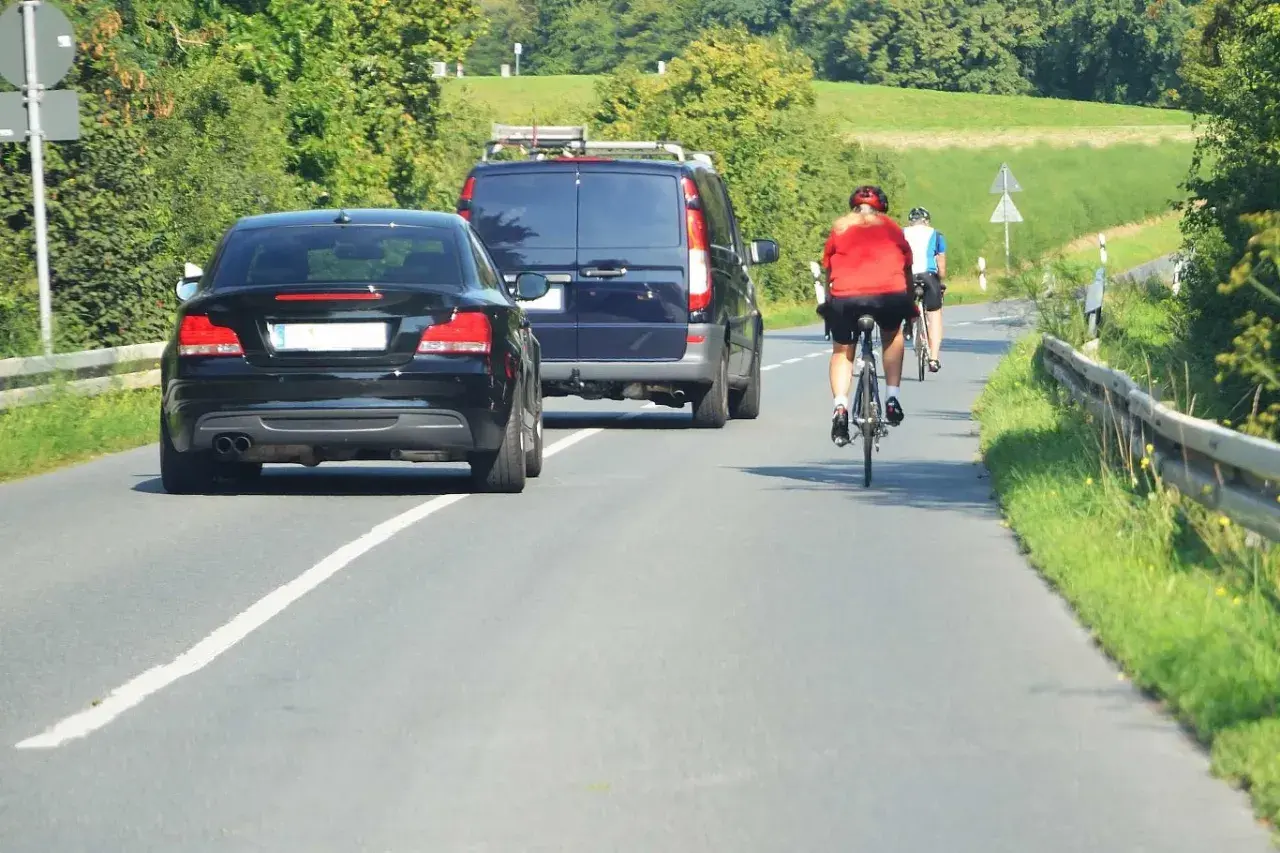 Rowerzyści na drodze, przestrzegający przepisy ruchu drogowego dla rowerzystów, mijani przez samochody i vana.