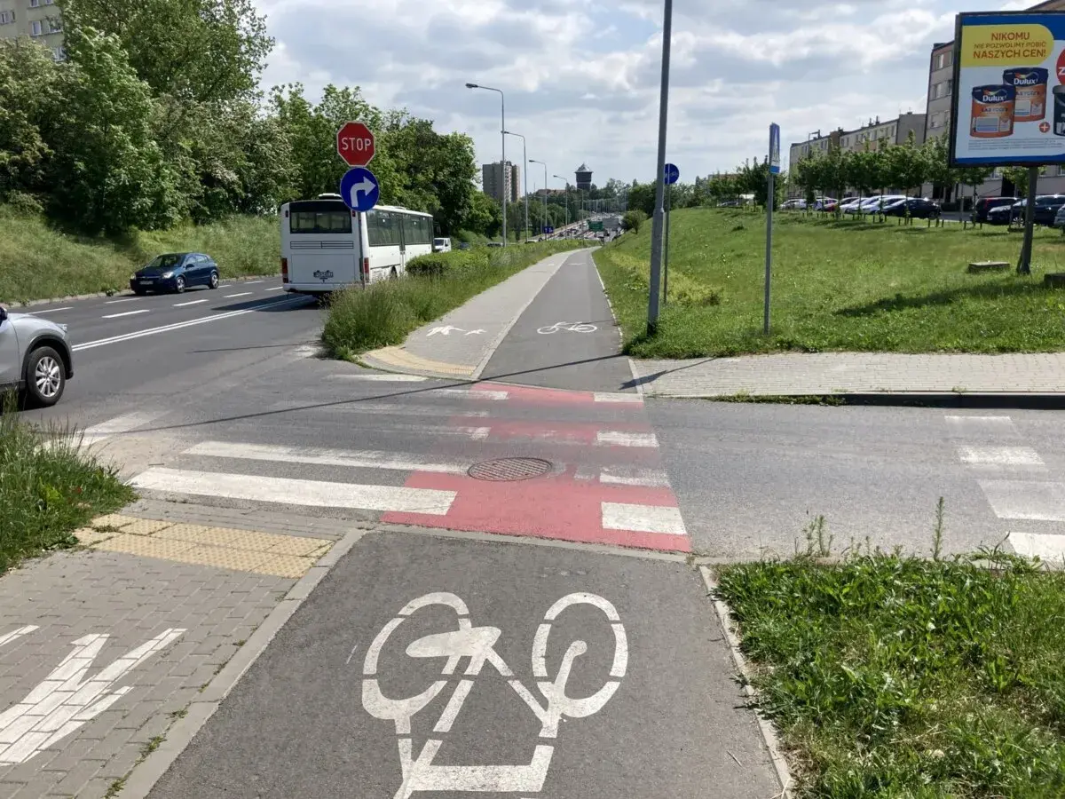 Przejście dla pieszych na ścieżce rowerowej, z oznaczeniem roweru. Autobus i samochody czekają na drodze.