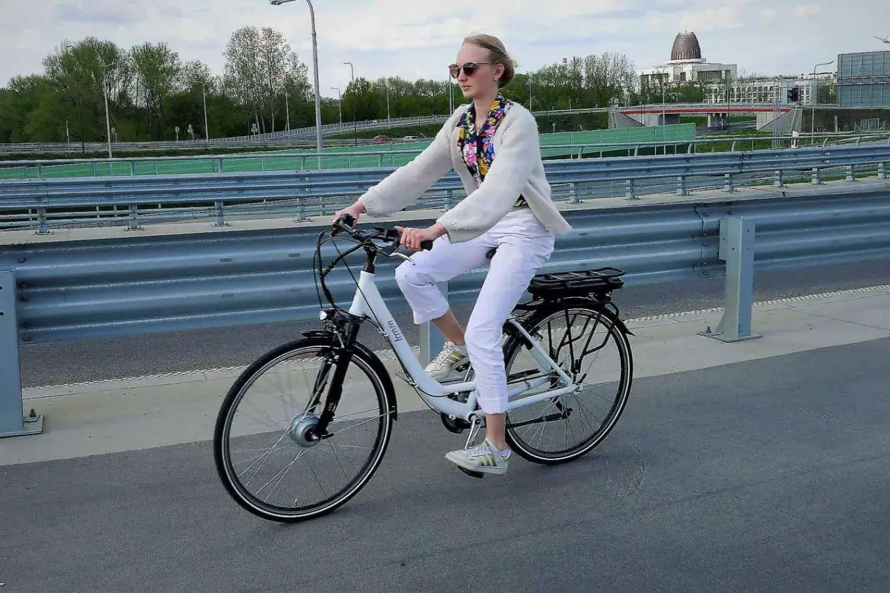 Kobieta w białych spodniach i sweterku jedzie na rowerze elektrycznym. W tle most i budynki.