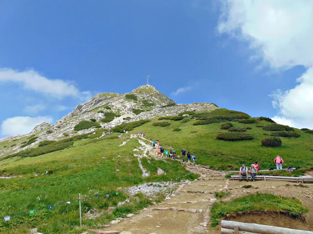 Tłum ludzi wspina się po kamienistym szlaku na Giewont. Widać krzyż na szczycie.