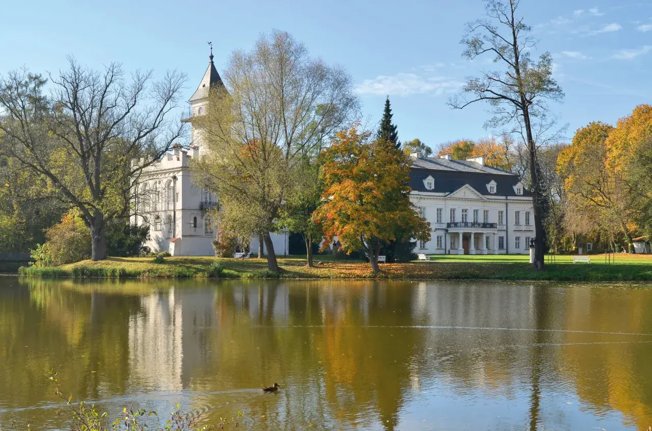Pałac w jesiennej scenerii, otoczony drzewami o barwnych liściach, odbija się w spokojnej tafli jeziora. W oddali widać fragment puszczy bolimowskiej.