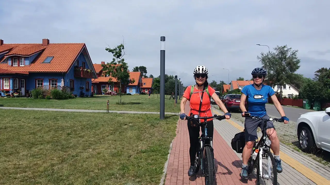 Dwie kobiety na rowerach mijają kolorowe domki nad rzeką Niemen.