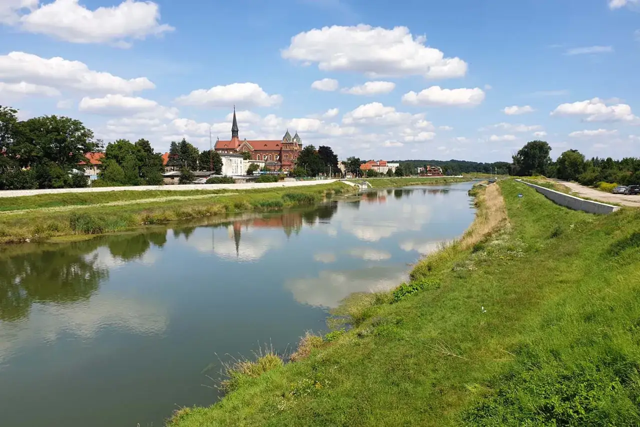Spokojna Nysa Kłodzka odbija chmury i miasto z kościołem. Zielone brzegi zapraszają na spacer.