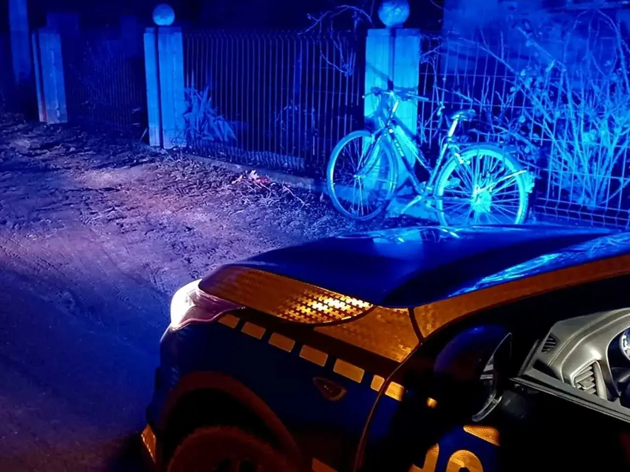 Nocna akcja policji. Czy można stracić prawo jazdy na rowerze? Rower stoi przy płocie, oświetlony niebieskimi światłami radiowozu.