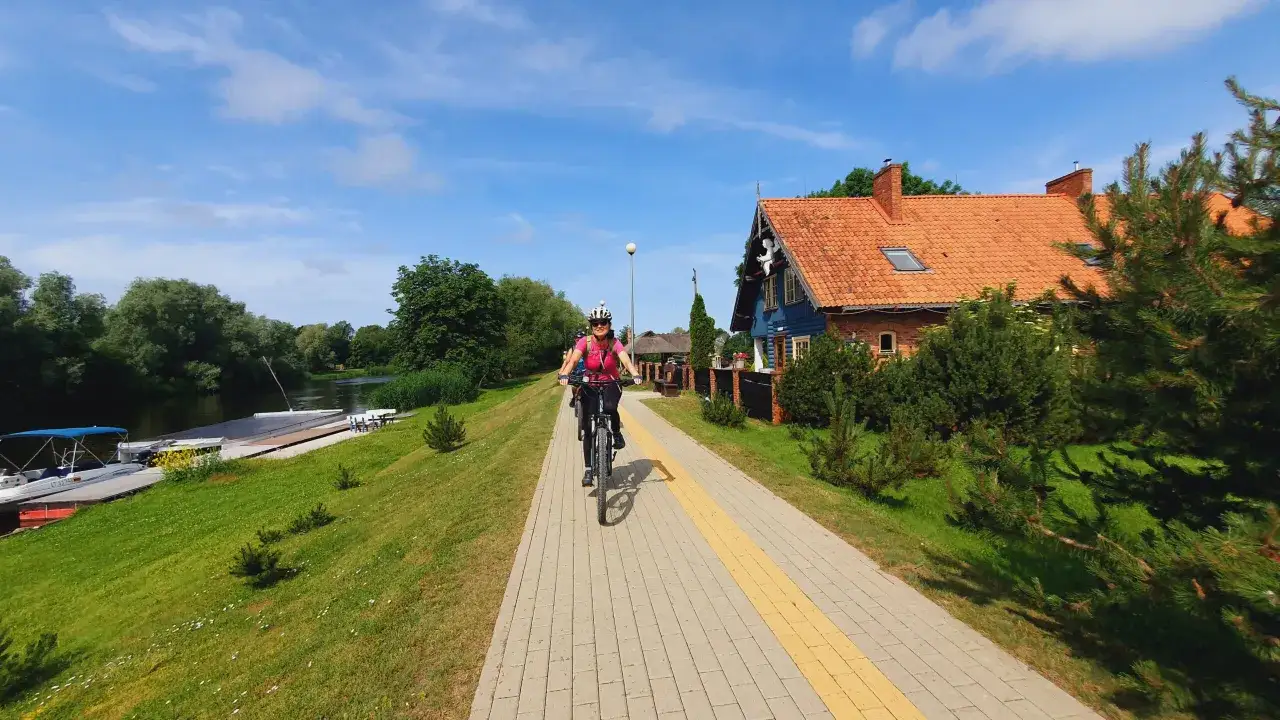Kobieta na rowerze jedzie ścieżką nad rzeką Niemen. W tle dom z czerwoną dachówką i zielone drzewa.