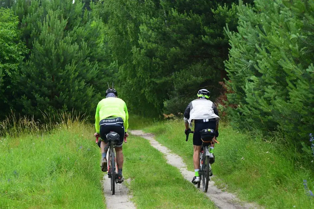 Dwóch rowerzystów jedzie ścieżką wśród drzew, w pobliżu rzeki Pilicy.