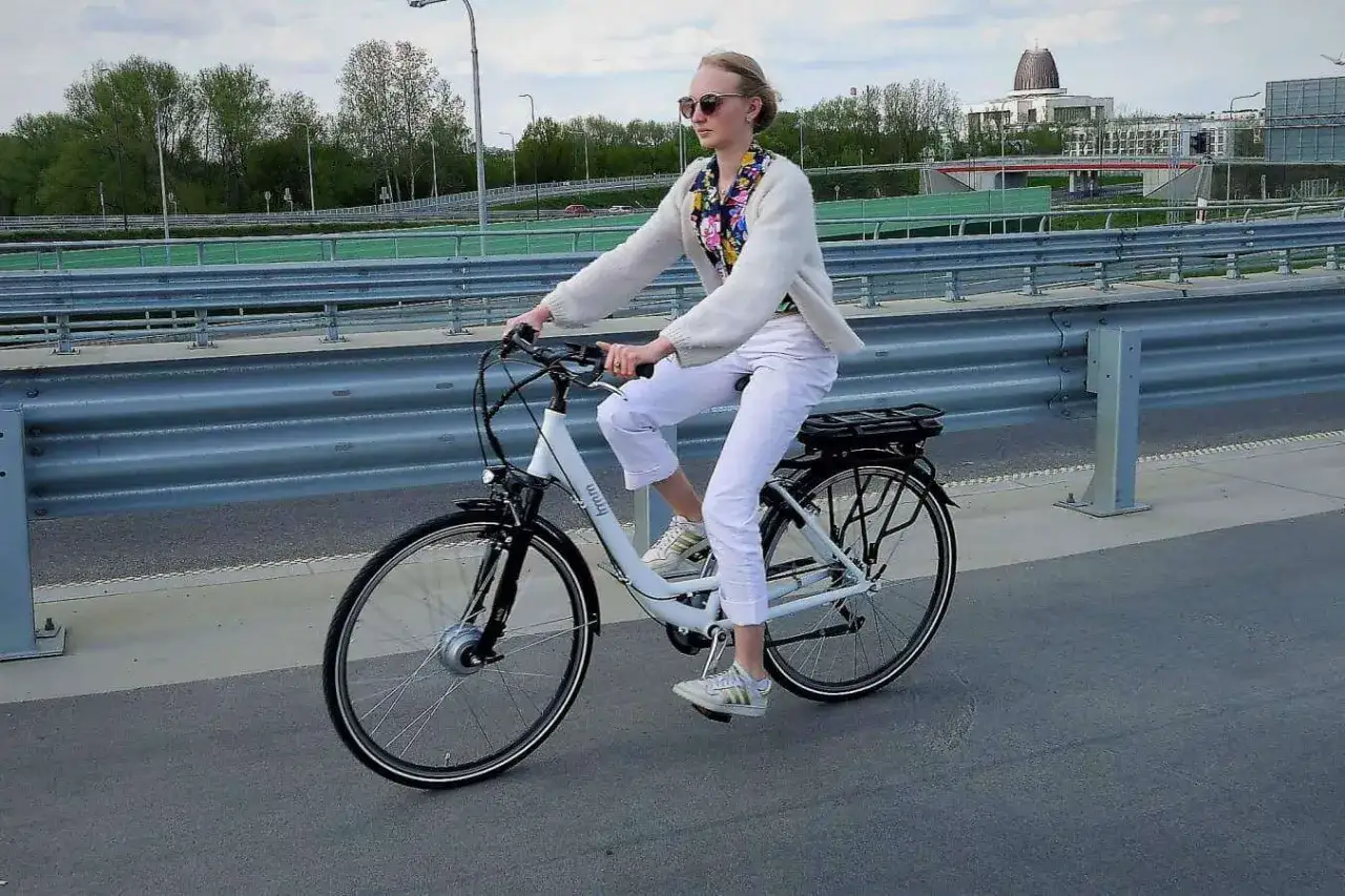Kobieta w białych spodniach i sweterku jedzie na rowerze elektrycznym. Przepisy dotyczące rowerów elektrycznych są coraz bardziej popularne.