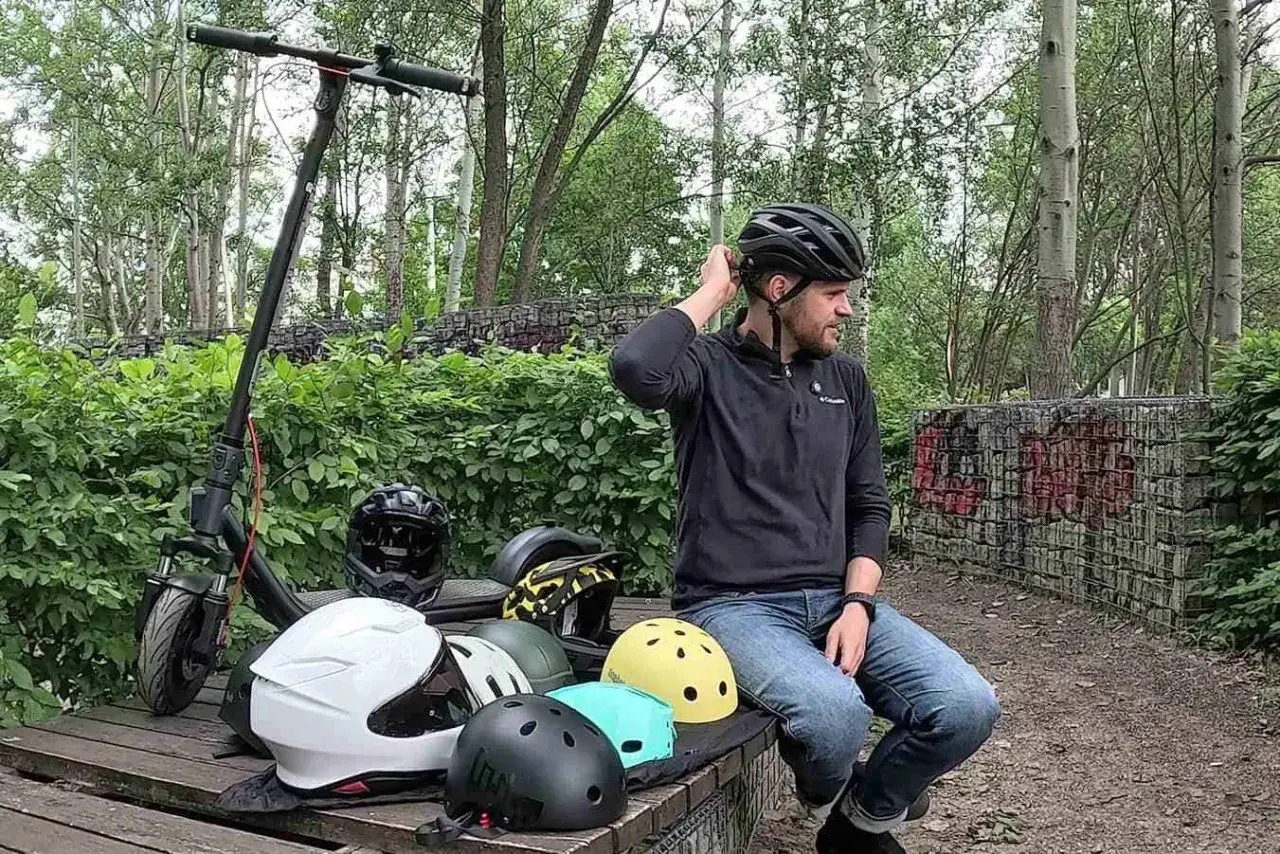 Mężczyzna w kasku na rowerze, otoczony różnymi kaskami i hulajnogą, przygotowuje się do bezpiecznej jazdy.